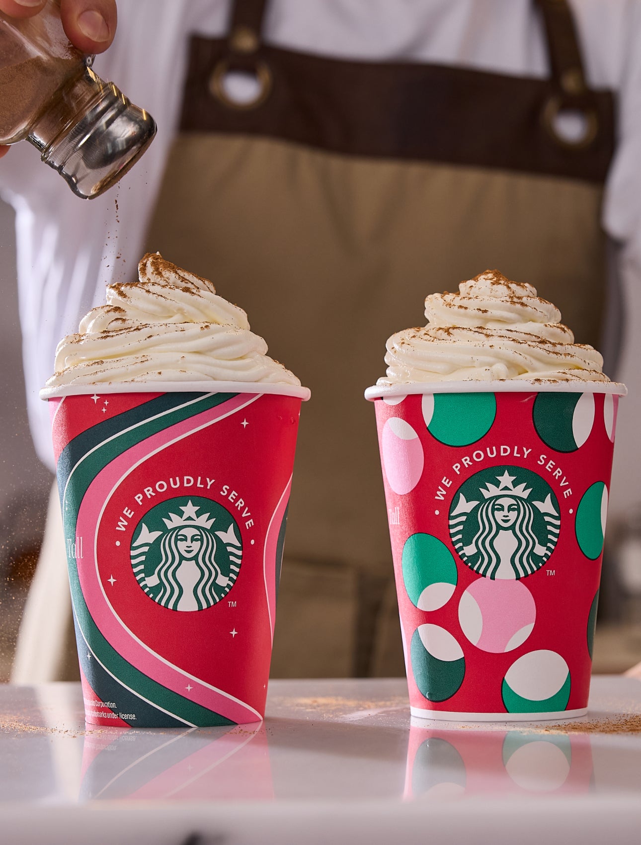 Twee Frappuccino® Gemixte Dranken in feestelijke We Proudly Serve Starbucks bekers, waarvan er één is bestrooid met kaneel. 