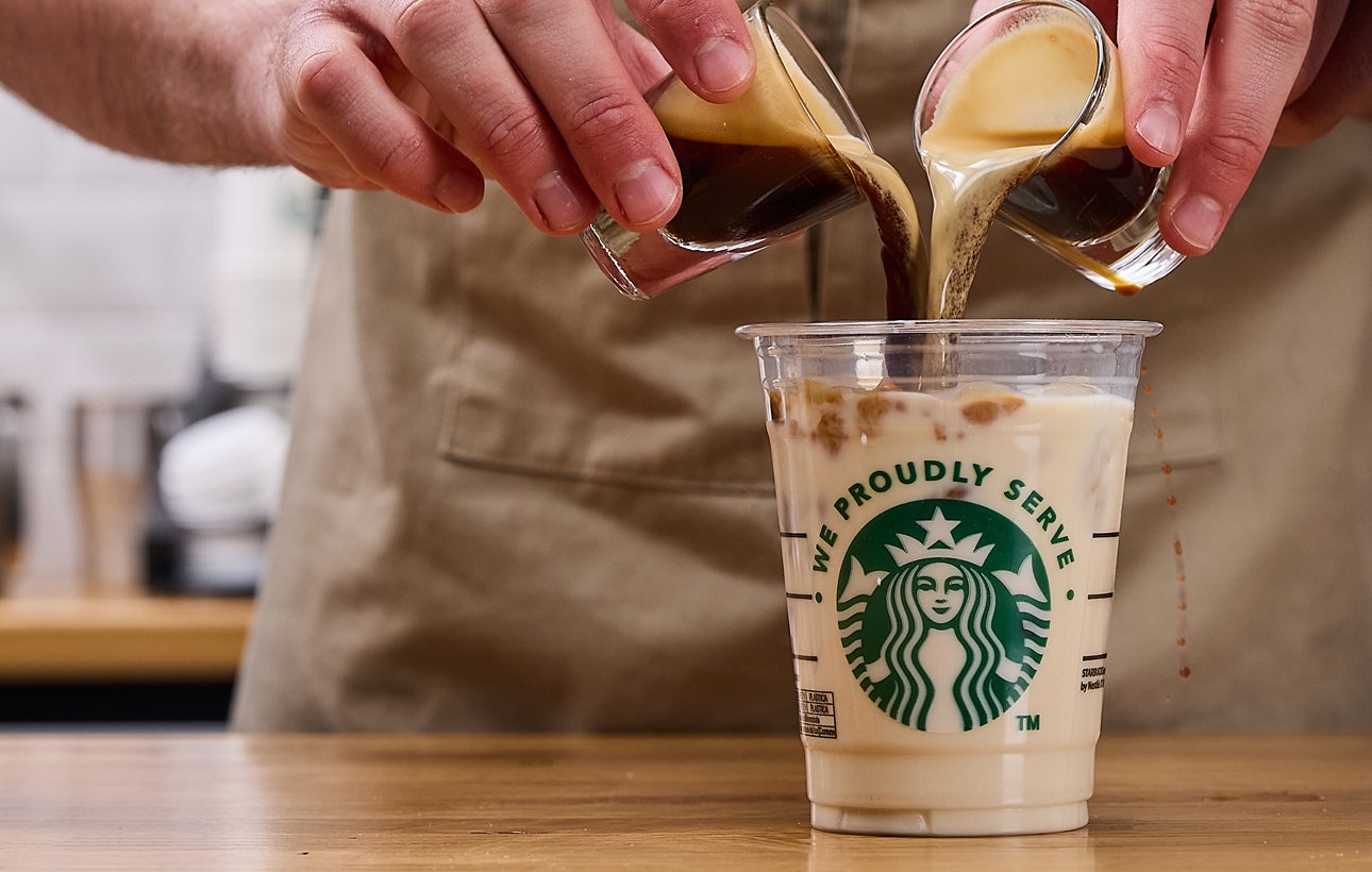 Een barista schenkt twee espressoshots tegelijk in een We Proudly Serve Starbucks beker gevuld met ijs en melk