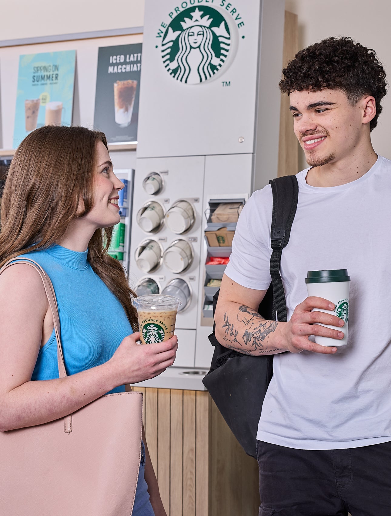 Een vrouw en een man staan voor een We Proudly Serve Starbucks zelfbedieningskoffiestation