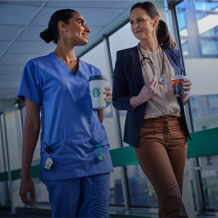 Twee zorgprofessionals lopen samen in een medische instelling, glimlachend en met een We Proudly Serve Starbucks drankje in d