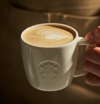 Een hand die een witte Starbucks mok vasthoudt, gevuld met een latte en versierd met hartvormige latte art.