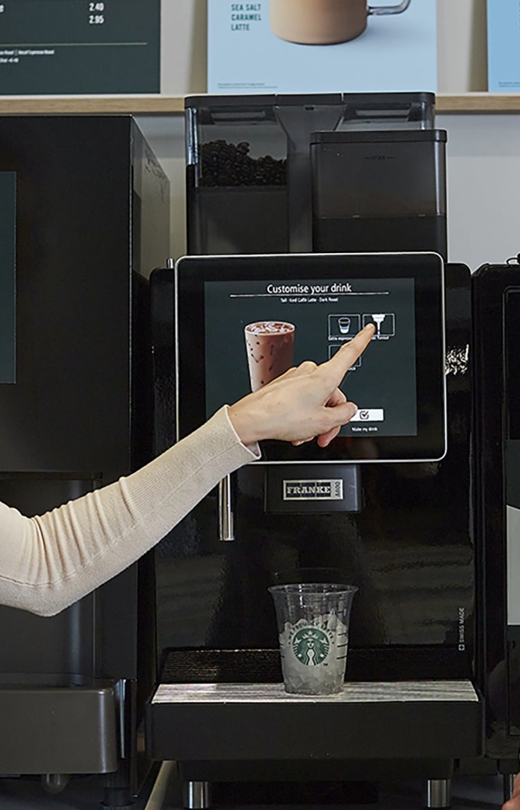 พนักงานบริษัท 2 คนกำลังกดกาแฟจากเครื่อง We Proudly Serve Starbucks รูปแบบบริการตนเอง